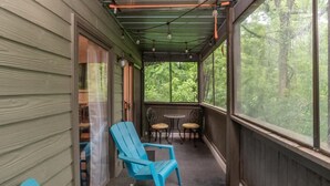 A covered porch with two blue chairs, a small table, and a view of the forest. - Woodland Escape Condo (Bloomington)