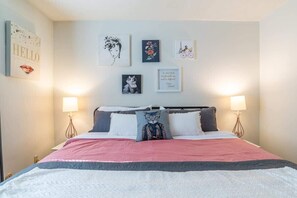 A neatly made bed with a pink and white bedspread, two bedside lamps, and a wall with various framed artworks. - Maple Row: Karma Vibe Like That (Bloomington)