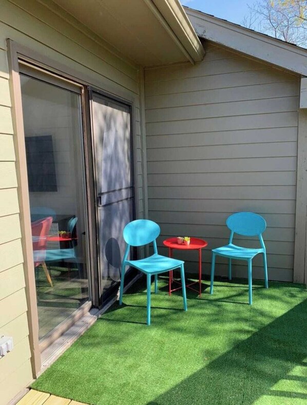 A small patio with artificial grass, two blue chairs, a red table, and a sliding glass door.