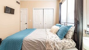 Une chambre à coucher avec un lit, un ventilateur de plafond, une télévision et une fenêtre avec des rideaux.