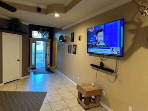 A living room with a television mounted on the wall, a small wooden stool, and a hallway leading to a door. - Spacious 4-bedroom house in Midland with AC (Midland)