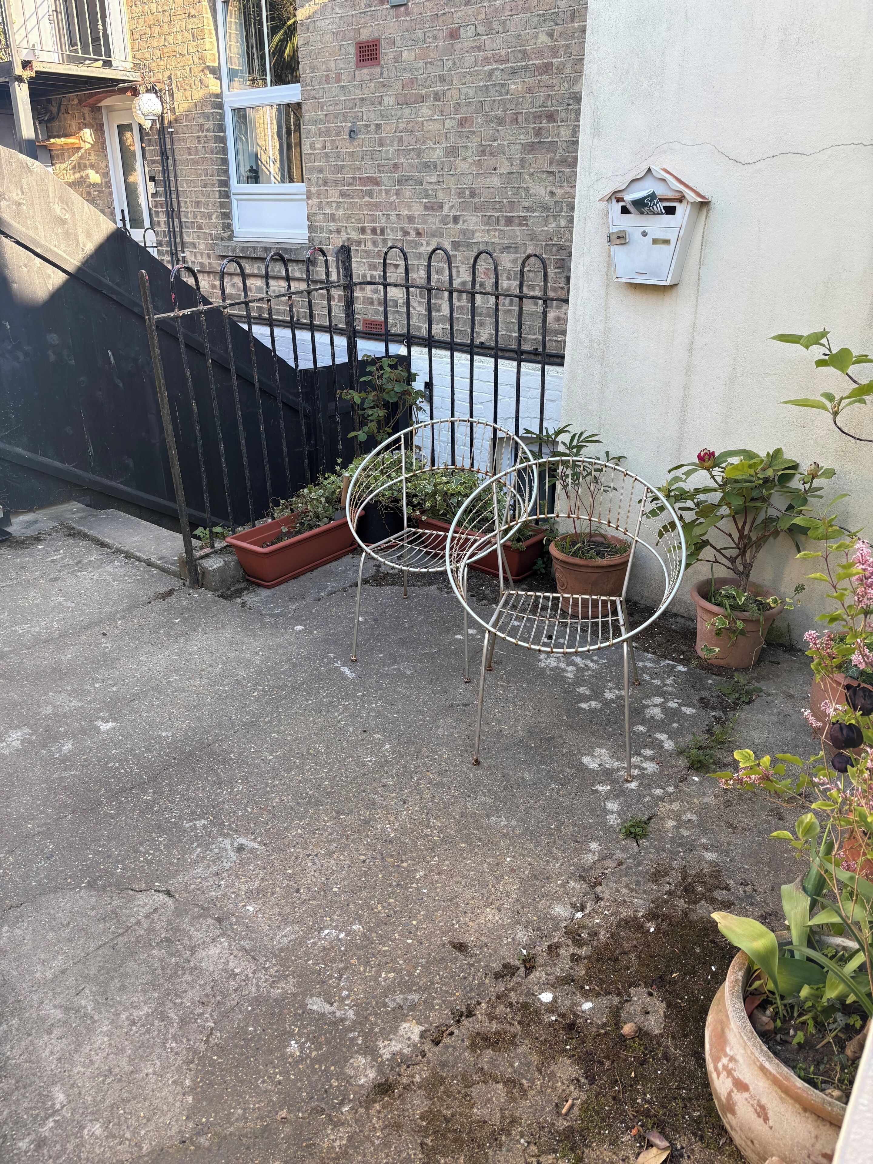 Une cour avec une chaise en métal, des plantes en pot, un mur de briques et un portail en métal.