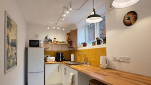 A compact kitchen with a refrigerator, microwave, toaster, and kettle. There is a window with a view of a brick wall, and a clock on the wall. - Stylish and peaceful 2 bedroom / 2 bathroom Oasis with a private Sun Courtyard (Saxmundham)