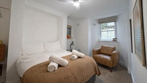 A bedroom with a bed, a brown chair, a ceiling fan, and a window. - Stylish and peaceful 2 bedroom / 2 bathroom Oasis with a private Sun Courtyard (Saxmundham)