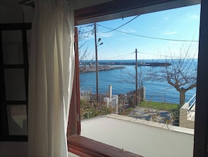 Blick aus einem Fenster mit Vorhang auf ein Gewässer, eine Anlegestelle und einen Baum.