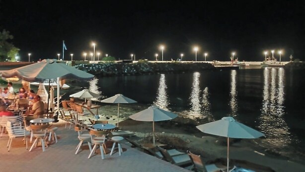 Un área de comedor frente al agua con mesas y sombrillas, un muelle con barcos y una masa de agua tranquila que refleja luces.