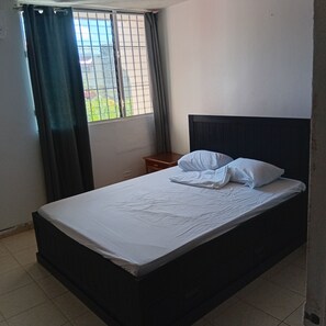 Uma cama de solteiro com cabeceira escura, uma mesinha de cabeceira e uma janela com cortinas.