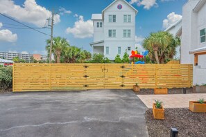 A gated driveway leading to a white building with a red and blue sign, surrounded by palm trees and a clear sky.