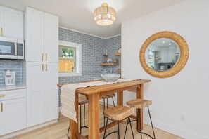 A kitchen with white cabinets, a wooden table, bar stools, a round mirror, and a hanging light fixture.