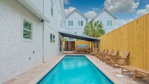 A rectangular swimming pool with a tiled deck, surrounded by a wooden fence and a white building with a covered patio area.