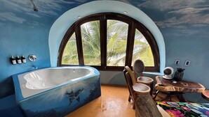 A room with a large bathtub, a wooden chair, a small table, and a window with a view of palm trees.