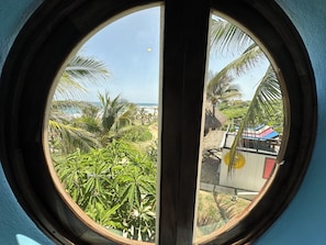 A round window with a view of a beach, palm trees, and a thatched-roof structure. - Two-story, two-bedroom oceanfront suite with private jacuzzis and full kitchen. (Santa Maria)