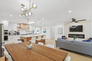 A modern kitchen with white cabinets, a central island, and a dining area with a wooden table and white chairs. - Dallas Home | Fire Pit · Spa Bath · Sleeps 11 (Dallas)