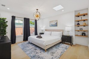 A modern bedroom with a large bed, bedside tables, a chandelier, a painting, and a view of the outside. - Dallas Home | Fire Pit · Spa Bath · Sleeps 11 (Dallas)