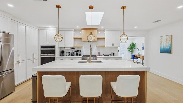 Una cocina moderna con armarios blancos, una isla central con taburetes y lámparas colgantes.