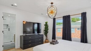 Un dormitorio con una cama, televisión, cómoda, planta y ventana con cortinas.