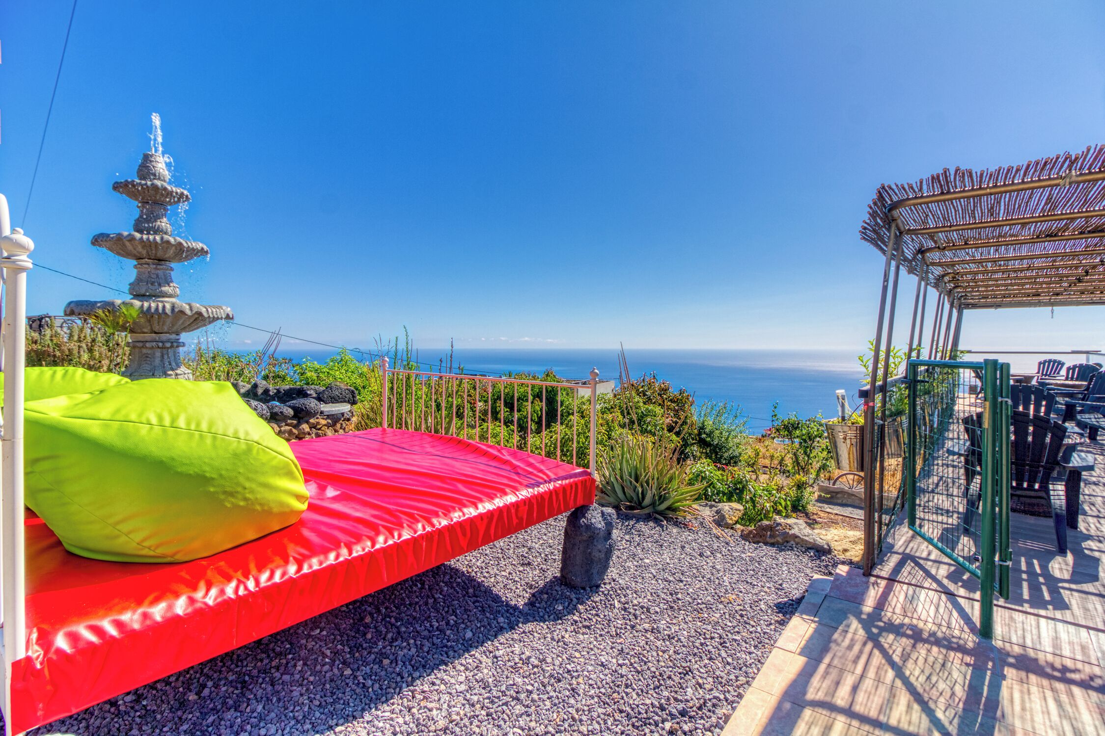 A red bed with pillows, a stone fountain, a view of the sea, and a covered seating area.