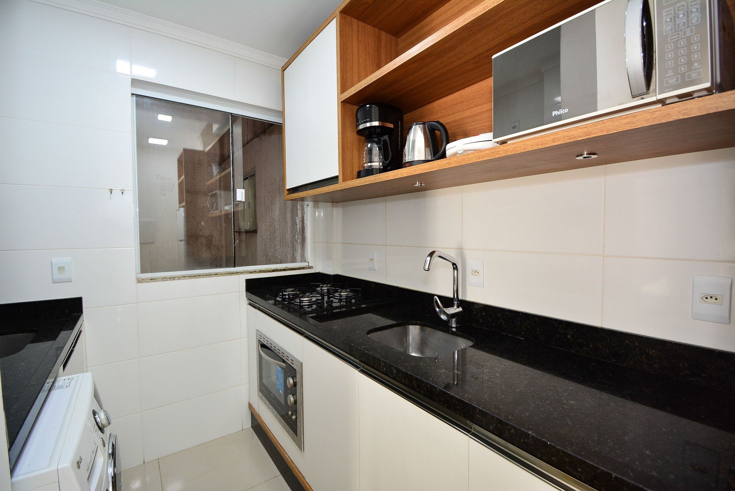 Una cocina con encimera negra, pileta de acero inoxidable y un microondas sobre una repisa de madera.