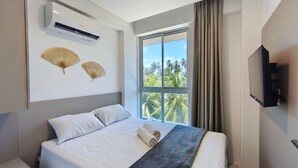 Hotellihuone, jossa on sänky, ilmastointilaite, kaksi seinätuuletinta, suuri ikkuna, josta on näkymä palmuille, ja taulutelevisio.