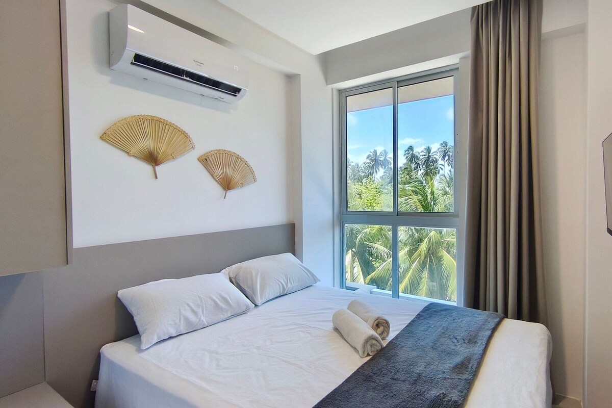 Ein Hotelzimmer mit Bett, Klimaanlage, zwei Wandventilatoren, einem großen Fenster mit Blick auf Palmen und einem Flachbildfernseher.