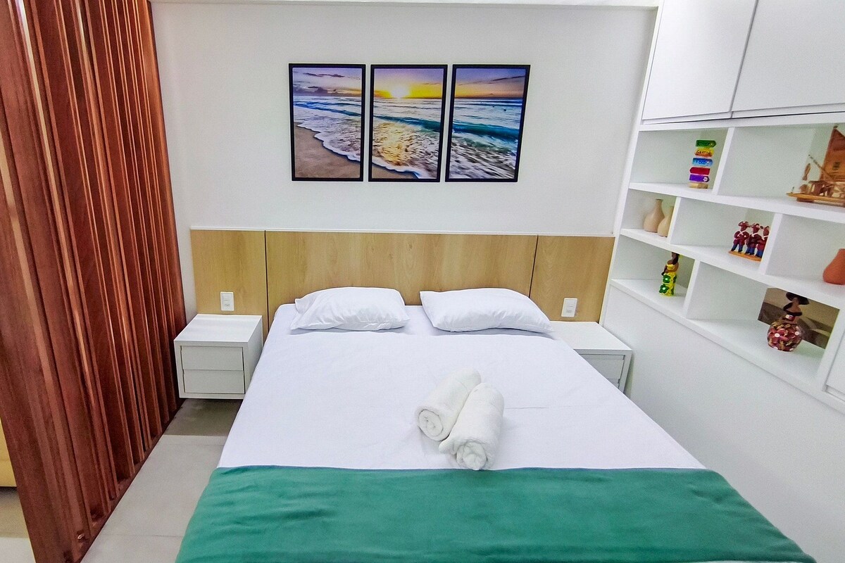 A hotel room with a bed, a nightstand, a bedside table, a sofa, a wooden headboard, a wall with a painting, and a shelf with decorative items.