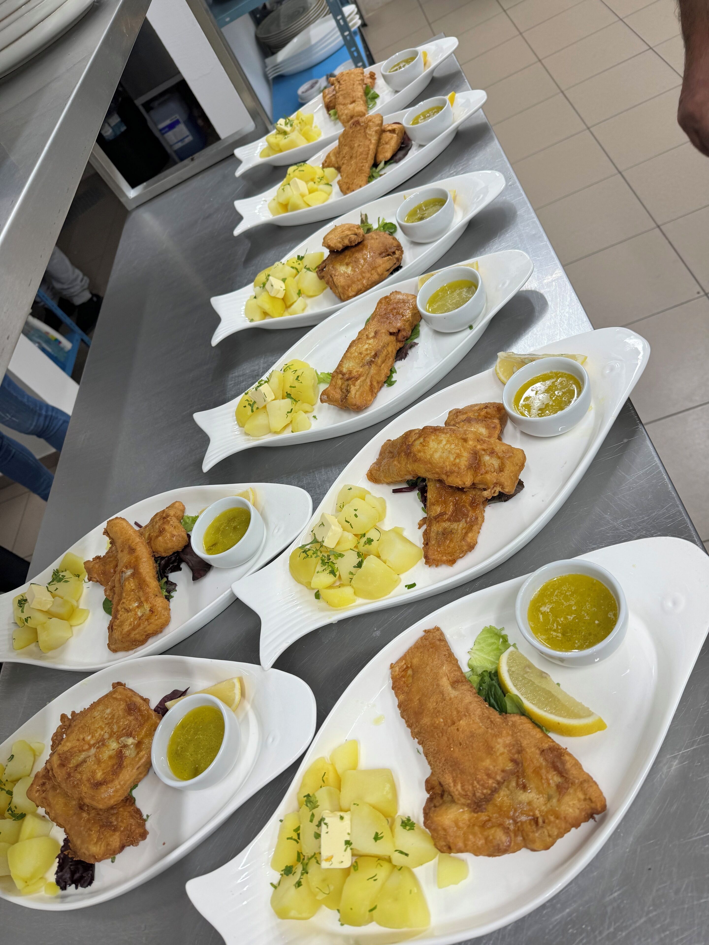 A row of white fish and potato dishes with lemon wedges and green sauce on a stainless steel counter.