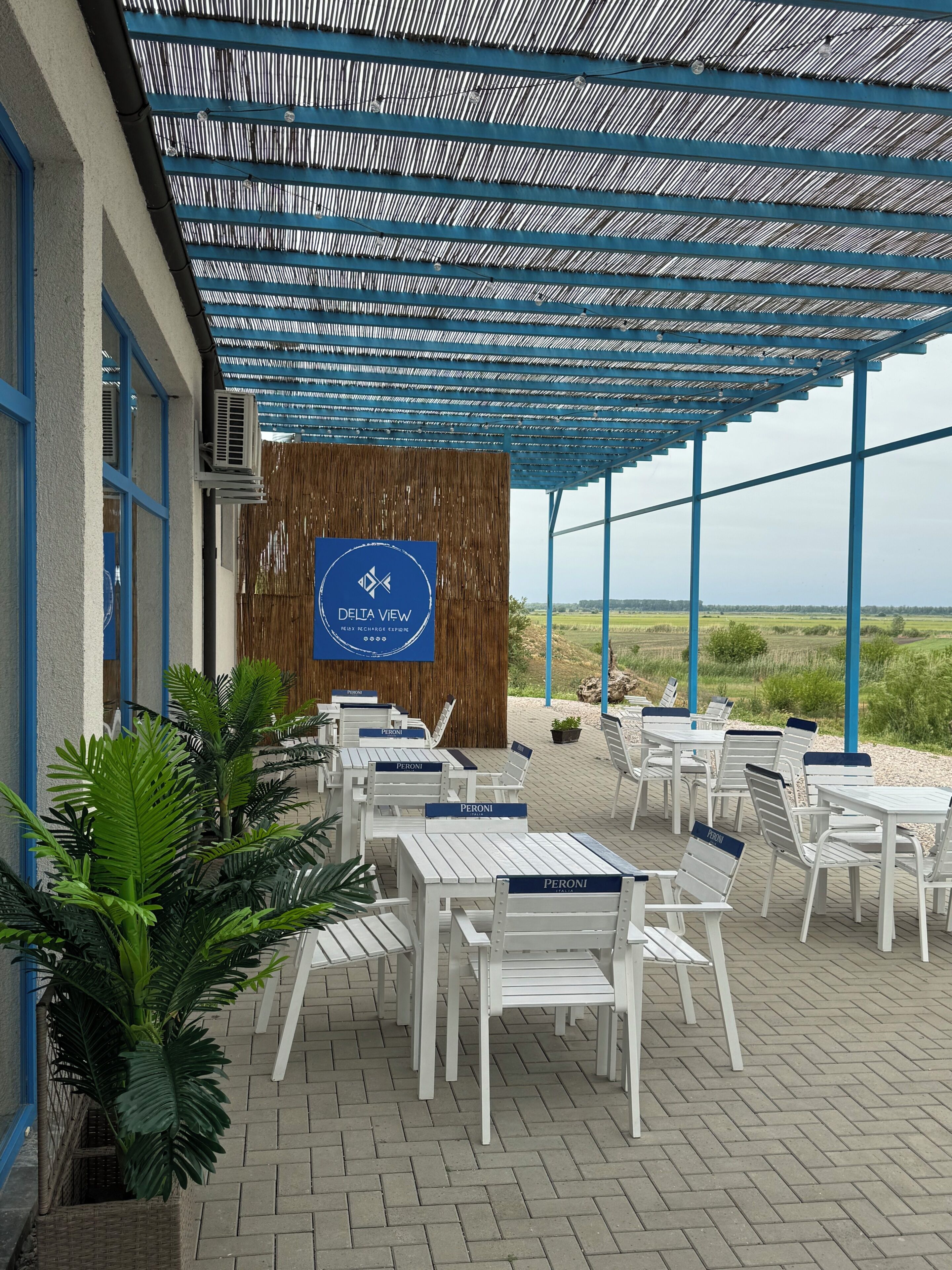 A covered outdoor seating area with tables and chairs, a wooden wall with a sign, and a view of the landscape.
