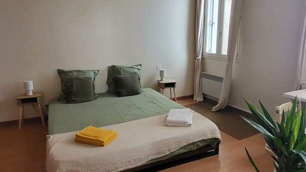 Ein Schlafzimmer mit Bett, Nachttischen, einer Lampe, einem Fenster mit Vorhängen und einer Zimmerpflanze.