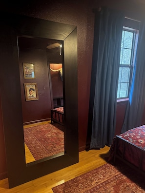 A room with a large mirror, a bed with a patterned bedspread, a wooden floor, and a window with curtains. - Honky Tonk Hideaway—where lake days turn into neon nights! (Canyon Lake)