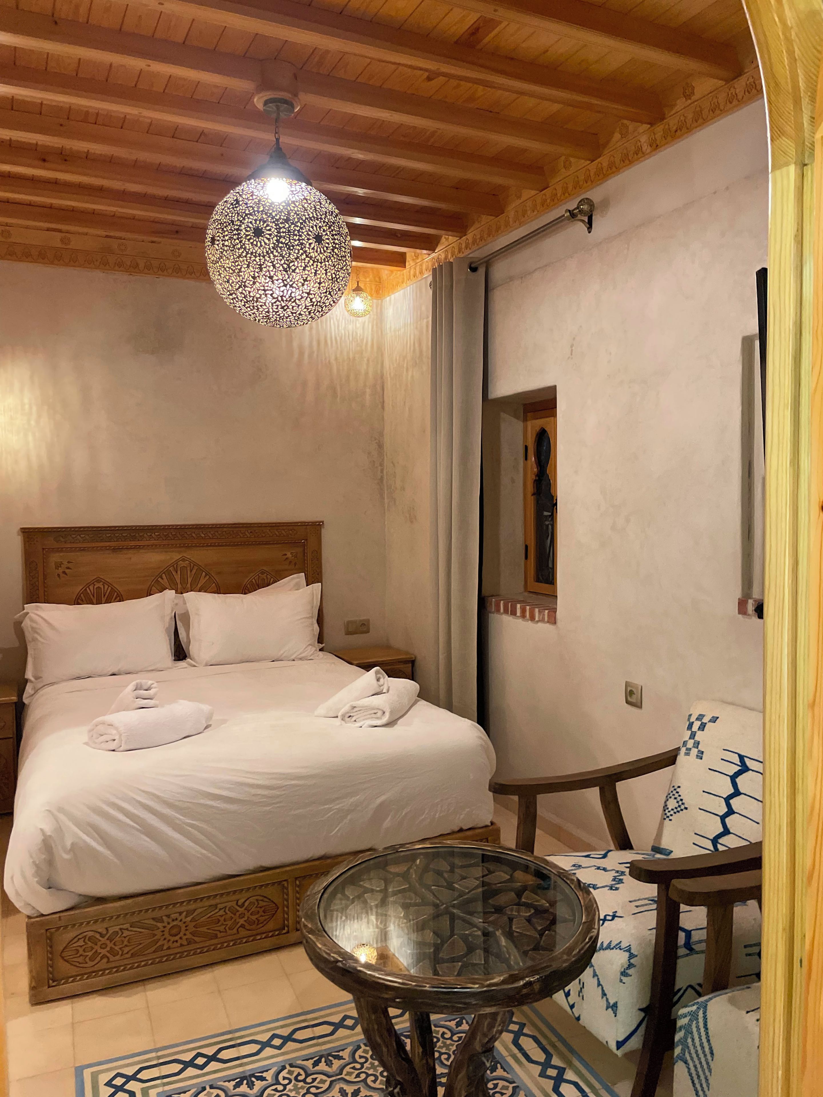 Habitación de hotel con techo de madera, una cama grande con ropa blanca, una mesa de vidrio y una silla estampada.