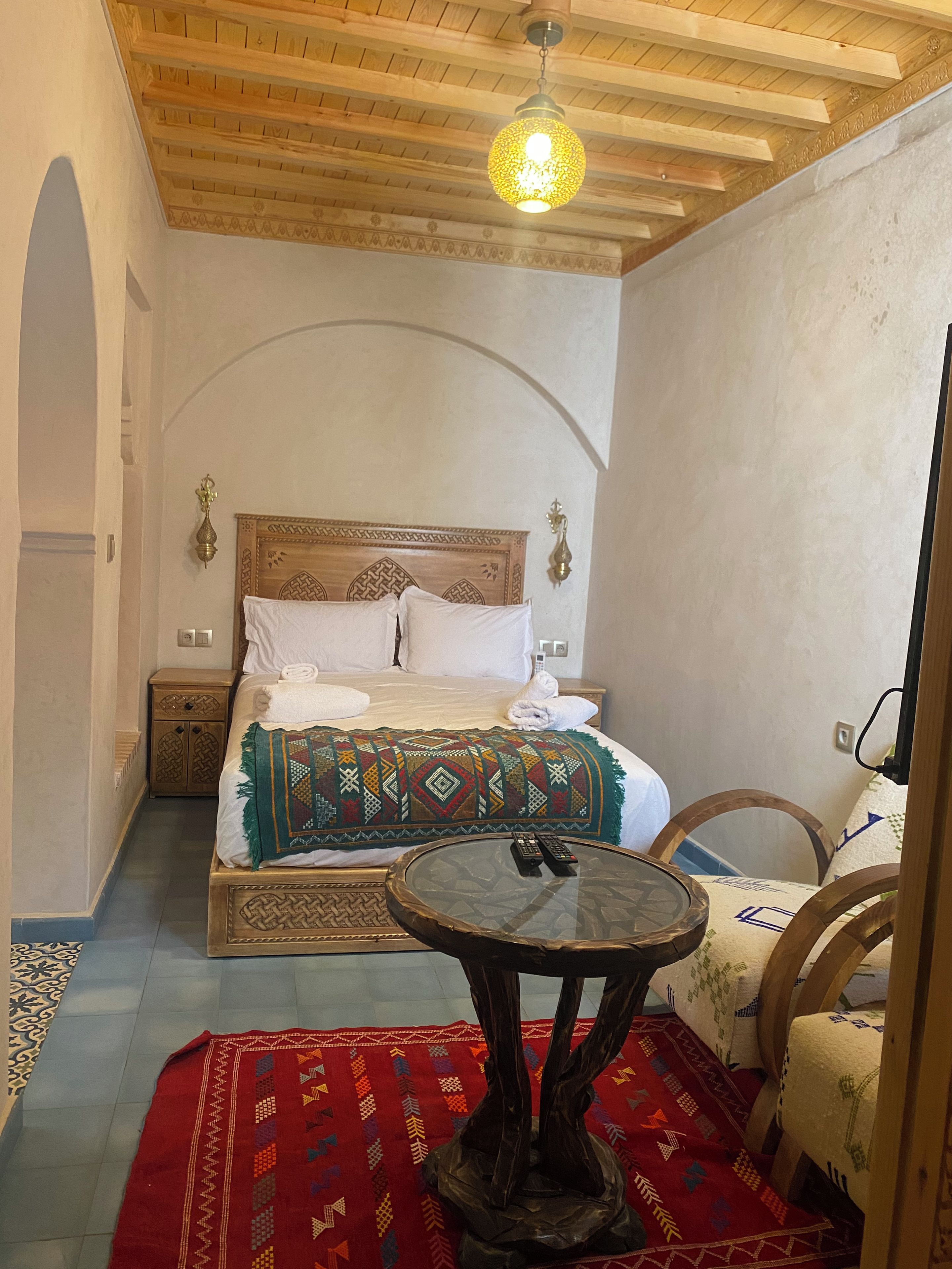 A bedroom with a wooden ceiling, a bed with a patterned bedspread, a round table with a remote control, and a red rug.