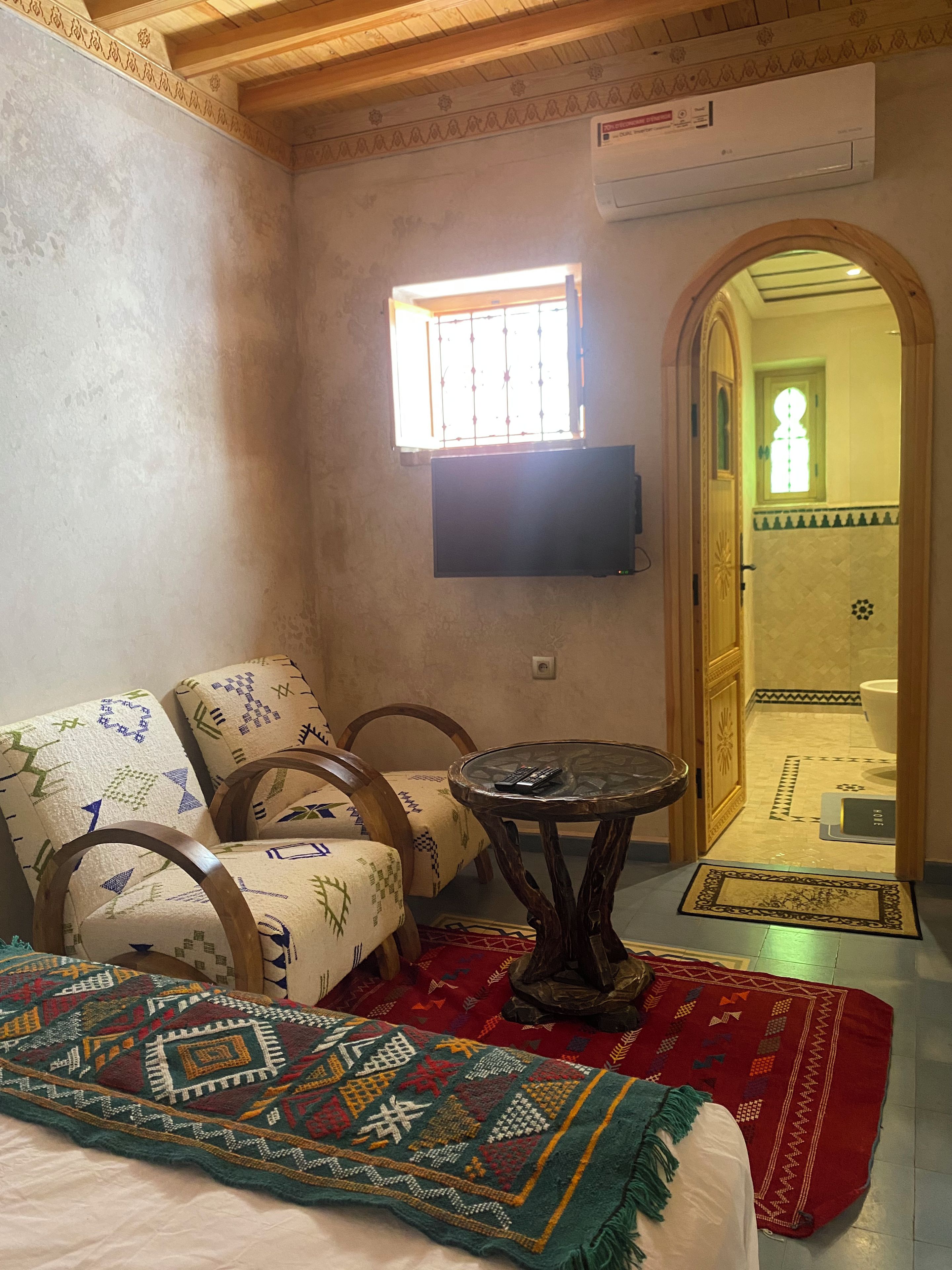A room with a bed, two chairs, a small table with a remote control, a window, and a door leading to a bathroom.