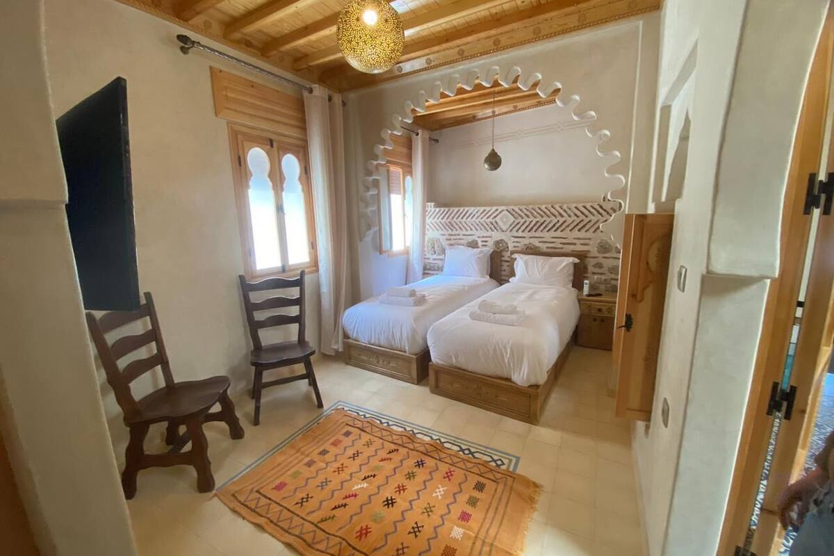 A bedroom with two beds, a wooden ceiling, a patterned rug, and a chair.