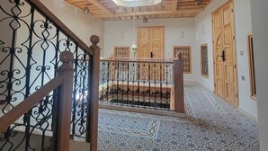 A hallway with a wooden ceiling, patterned floor tiles, a staircase with a wrought iron railing, and wooden doors.