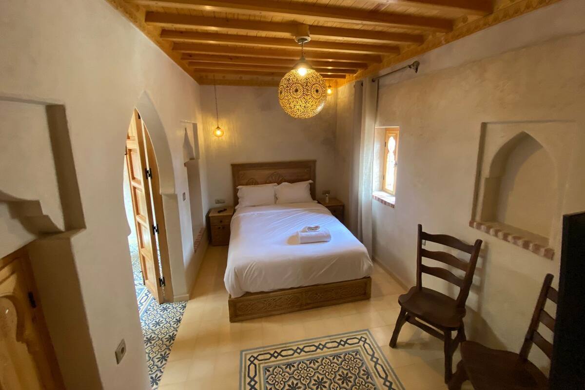 A bedroom with a wooden ceiling, a bed with white linens, a nightstand, a chair, and a patterned rug.