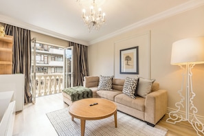 Ein Wohnzimmer mit einer Couch, einem runden Couchtisch, einer Chandelieber, einer Lampe und einem Balkon mit Blick auf Gebäude.