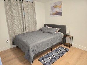 Een slaapkamer met een bed, gordijnen, een nachtkastje met een lamp, een vloerkleed en een kunstwerk aan de muur.