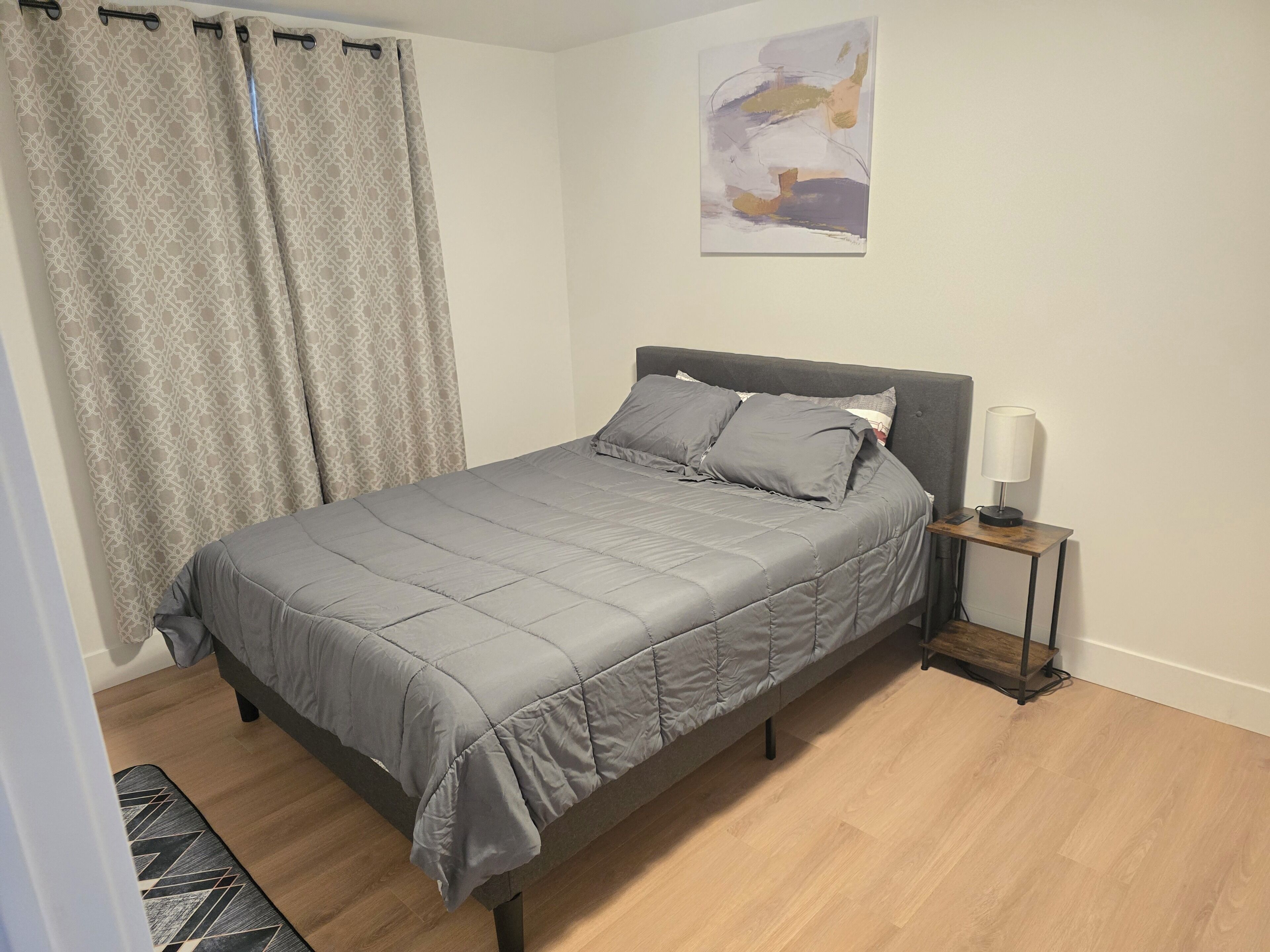 A bedroom with a bed, curtains, a bedside table with a lamp, and a wall art piece.