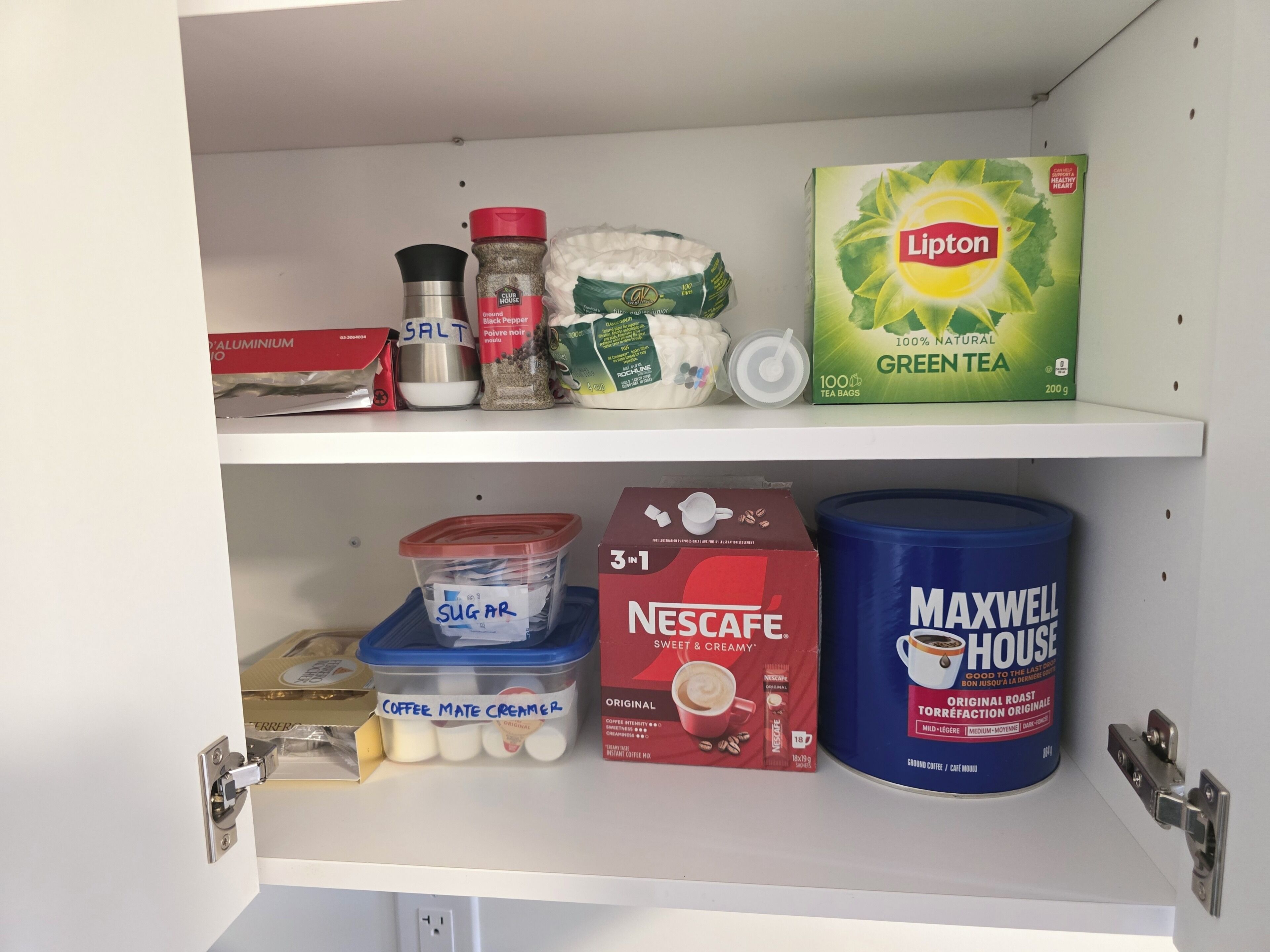 Een keukenkastje met verschillende items, waaronder Lipton groene theezakjes, een suikerpot, een melkpak voor koffie, een doos Nescafe sweet & creamy koffie en een Maxwell House koffiebus.
