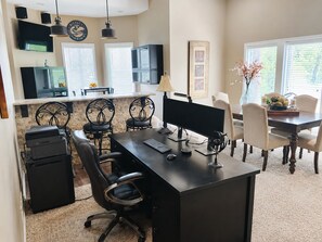 A home office with a desk, computer, printer, and chairs. There are windows with blinds, a dining table with chairs, and a kitchen area with a refrigerator.