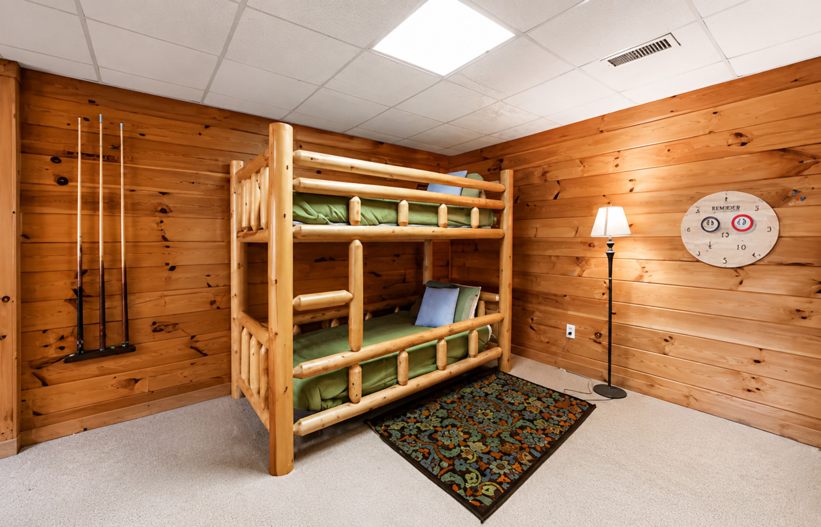 Quarto com beliches, paredes de madeira, colcha verde, tapete estampado, relógio de parede e abajur.