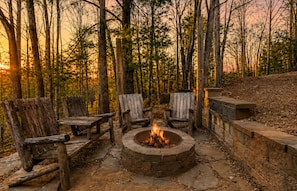 Ein kreisförmiges Lagerfeuer, umgeben von Holzstühlen in einer Waldlandschaft.