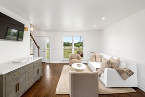 A spacious living room with a white sofa, a beige armchair, and a coffee table. A large window offers a view of the outdoors.