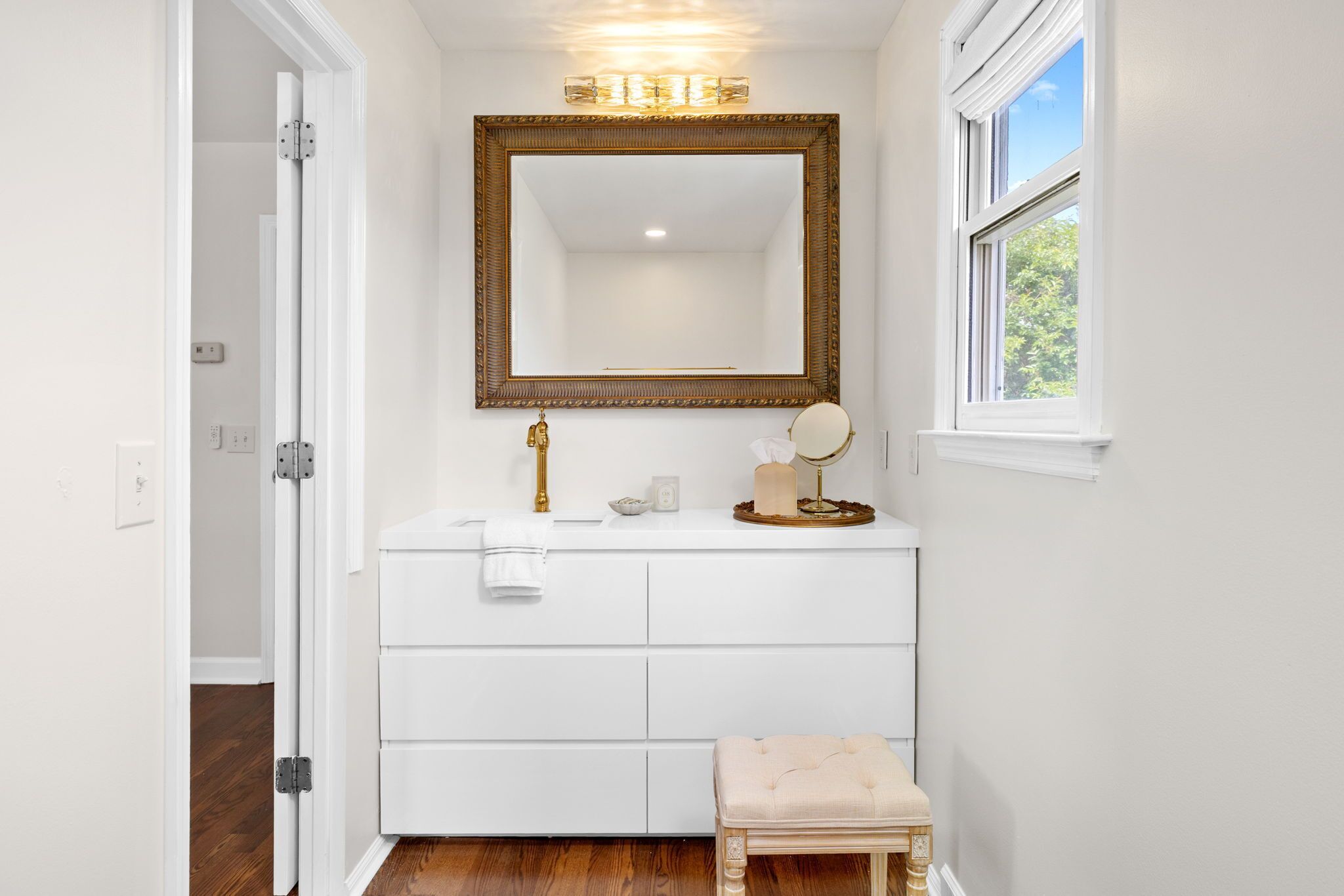 A bathroom with a white vanity, a large mirror, a window, and a door leading to another room.