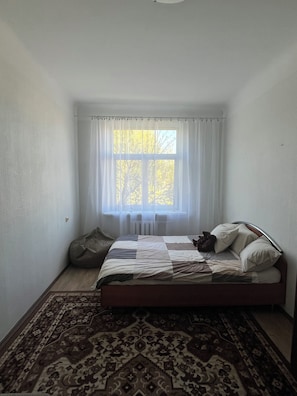 Un dormitori amb un llit, una finestra amb cortines, una catifa amb dibuixos i un radiador.
