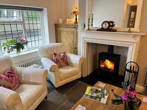 A cozy living room with two armchairs, a fireplace, a radiator, and a window with flowers. - Bell Cottage, a period cottage between the North Yorkshire Moors & Dales (Brompton)