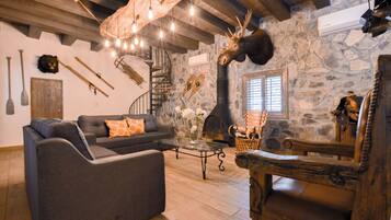 A rustic interior with a stone wall, wooden ceiling beams, a fireplace, a sofa, and a dining table.