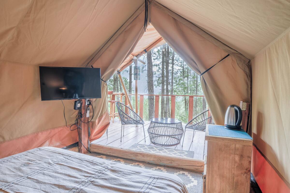 A tent with a bed, a TV, a table with chairs, and a view of trees.