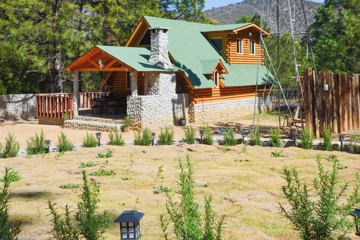 A rustic cabin with a stone chimney, a wooden deck, and a windmill in the background.