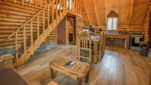 Interior de una cabaña de troncos con escalera de madera, comedor, cocina y chimenea de piedra.